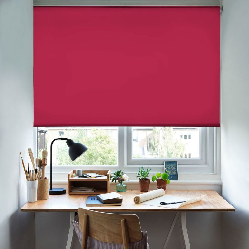 Fuchsia roller blind, fully lowered across a double window with hem; blocking most light while a narrow bottom gap admits daylight — above a wooden desk with lamp, plants, stationery.