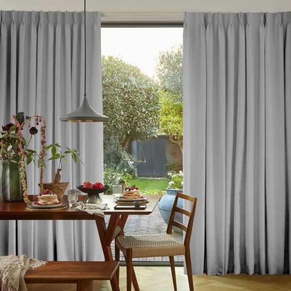 Object: light-gray, floor-to-ceiling pinch-pleat curtains parting at center; Action: framing a bright opening and admitting diffuse daylight; Context: wooden dining table set for breakfast, pendant lamp, garden view.