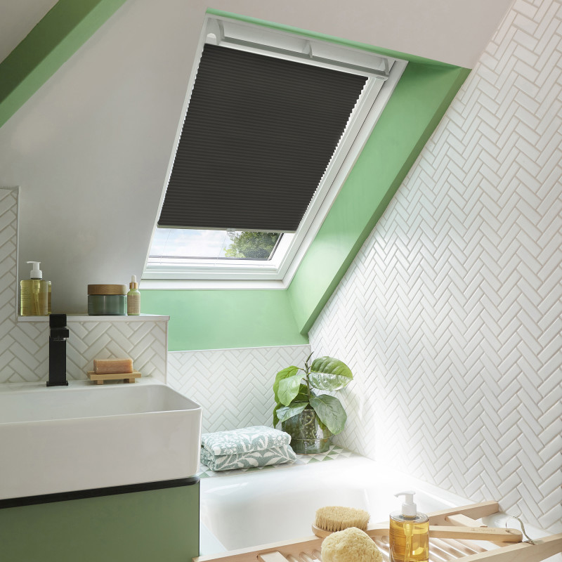 Black pleated skylight blind, partially lowered across a white roof window, blocking most light; attic bathroom with mint-green sloped walls, white herringbone tiles, sink, bathtub, plant and bath tray.