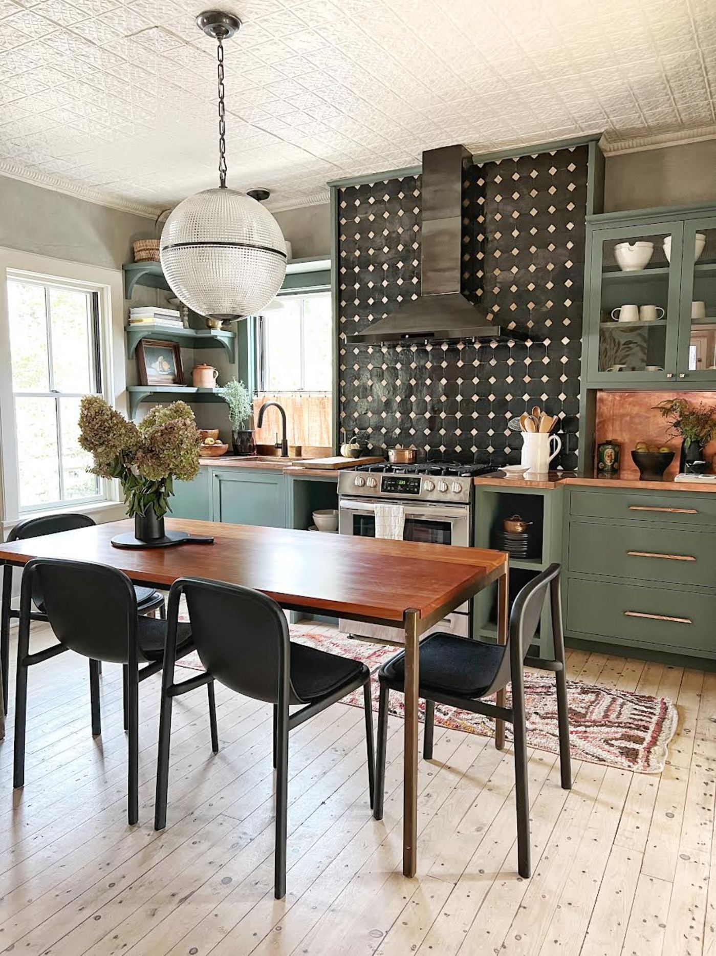 a kitchen with green cabinets, black and white tiled backsplash and a wooden table and chairs.