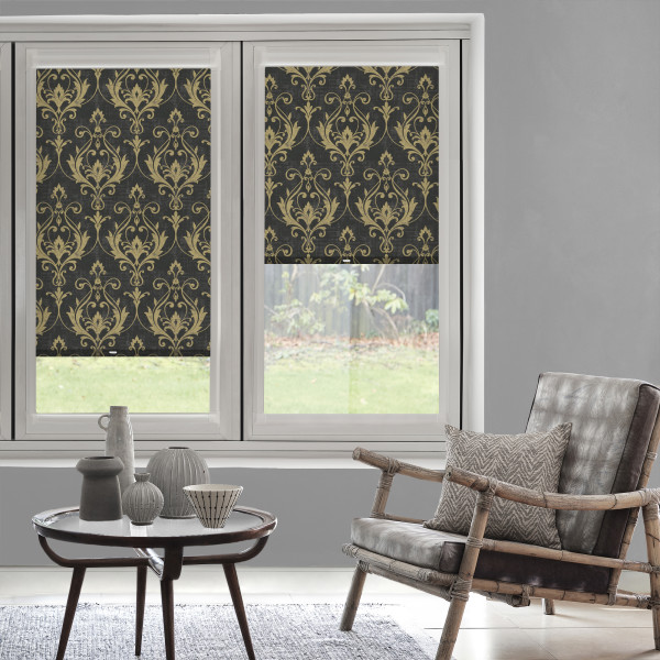 Patterned roller blinds (gold damask on black) lowered over a two-pane window, partially blocking light; modern living room with gray walls, rattan armchair, small table and backyard lawn view.