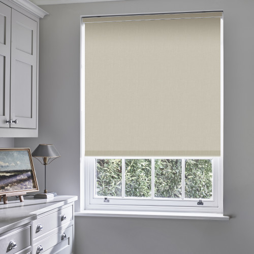 Beige roller blind lowered nearly fully, filtering light and covering the upper sash; gray-painted room with white cabinetry, lamp and painting, sash window shows leafy hedge outside.