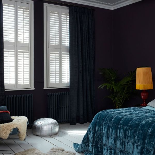 White plantation shutters on two tall windows, paired with dark floor-length curtains drawn to one side; shutters closed, filtering sunlight into a dim, moody bedroom with velvet bedspread, lamp, plant.