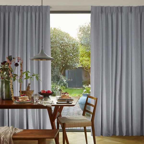 Floor-length grey pleated curtains, parted centrally, framing a glass door; filtering daylight into a dining nook with wooden table, pendant lamp, pancakes on plates, chairs, and a leafy garden view.