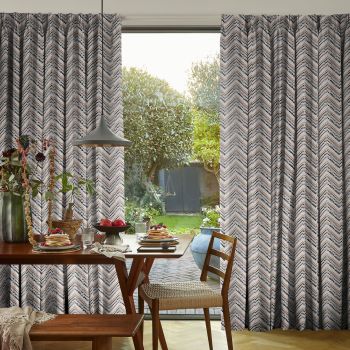 Patterned pleated curtains, floor-length with dotted chevron print, pulled partly aside to reveal a glass sliding door; dining table set for breakfast under a pendant lamp, sunlit garden beyond.