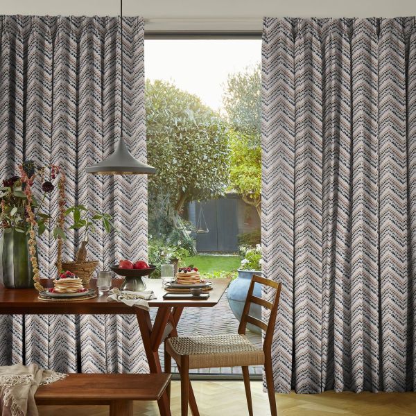 Patterned pleated curtains, floor-length with dotted chevron print, pulled partly aside to reveal a glass sliding door; dining table set for breakfast under a pendant lamp, sunlit garden beyond.