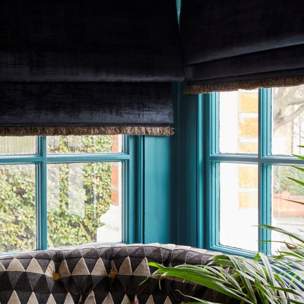 Dark velvet Roman blinds with fringed trim, partially lowered and dimming daylight, over turquoise-painted mullioned corner windows in a room with patterned seating, a plant; brick and garden visible outside.