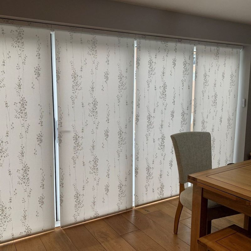 Patterned fabric panel blinds covering sliding glass doors, drawn closed with narrow gaps between panels; filtering soft daylight into a dining room with hardwood floor, wooden table and upholstered chair.