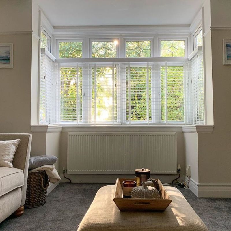 White slatted horizontal blinds covering a threeâ€‘pane bay window with decorative transom panels, partially open and filtering warm sunlight and leafy garden view into a neutral-toned living room with ottoman.