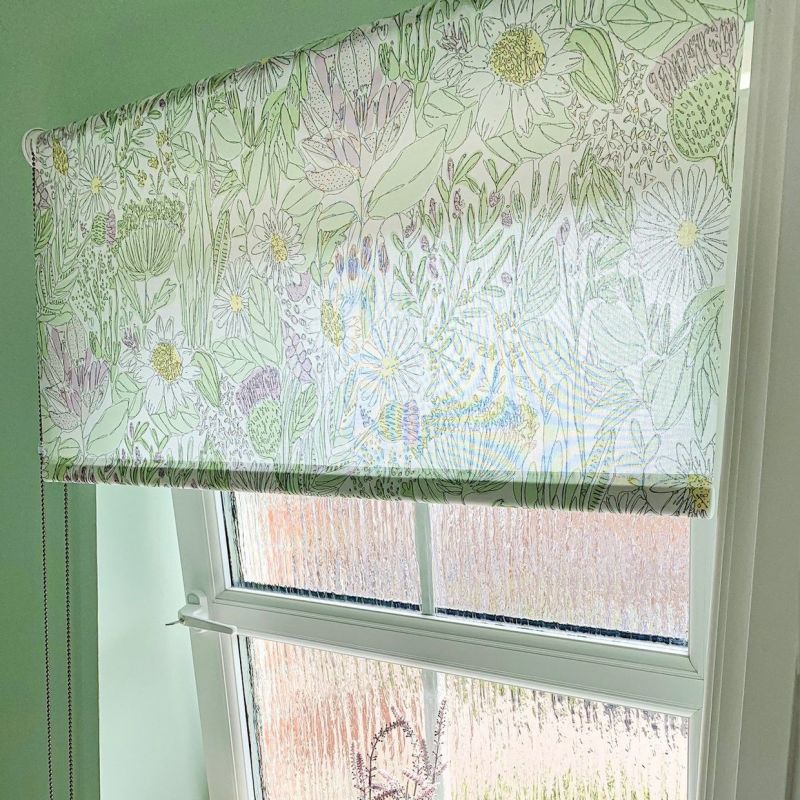 Floral roller blind with pastel green line-drawn flowers, lowered to mid-window, filtering bright daylight; mounted in a pale green-walled room over a frosted glazed sash window, metal pull chain visible.