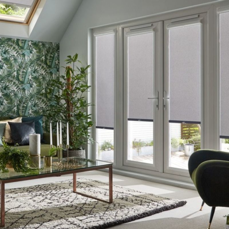 Grey fabric roller blinds on four tall French-door panels, lowered to mid-height and filtering daylight — in a living room with tropical wallpaper, skylight, plants, coffee table and patterned rug.