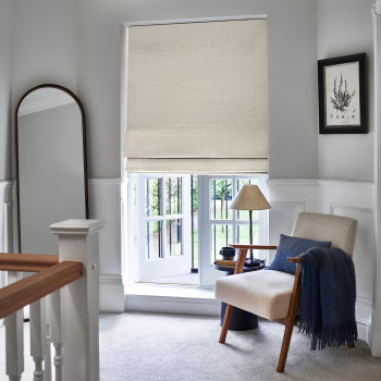 Beige dotted Roman shade lowered over window, softly folded at bottom, filtering daylight into a bright upstairs landing nook with upholstered armchair, lamp, full-length mirror and garden view.