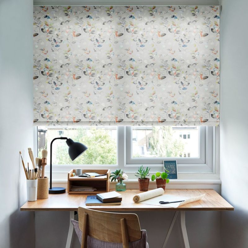 Patterned roller blind, lowered across the window, with a pale watercolor leaf-and-flower print, softly filtering daylight; above a tidy home desk with lamp, brushes, notebooks and potted plants.