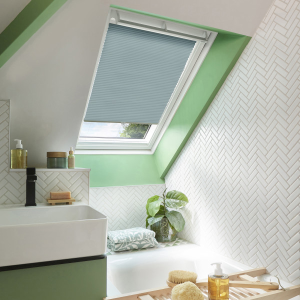 Light-blue pleated skylight blind, lowered halfway and filtering soft daylight; in a sloped attic bathroom with green-painted trim, white herringbone tiles, a basin, bathtub, and a potted plant.