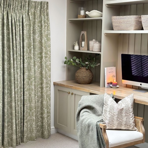 Pleated sage-green floral curtains, full-length and drawn closed, gently filtering daylight; beside a neutral built-in desk with wicker vase, iMac, lit candle, basket shelves, and a chair with throw.
Text: WHITE