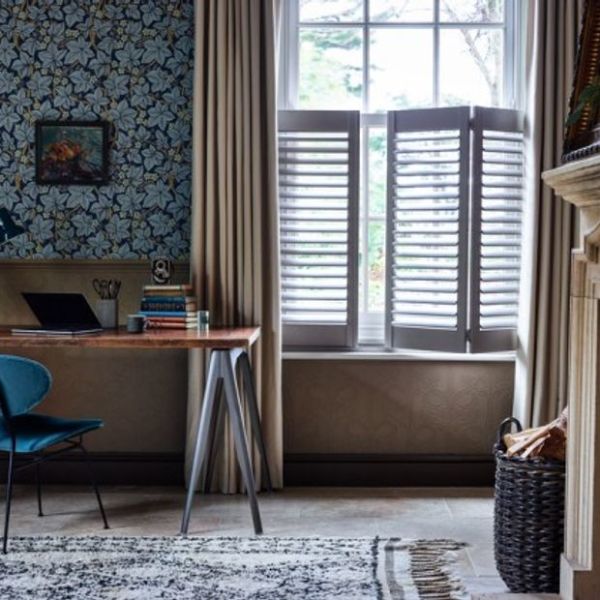 Beige floor-length curtains frame a tall window; white plantation shutters cover the lower sash with tilted horizontal slats, partially open and filtering daylight into a cozy study—desk and patterned wallpaper.