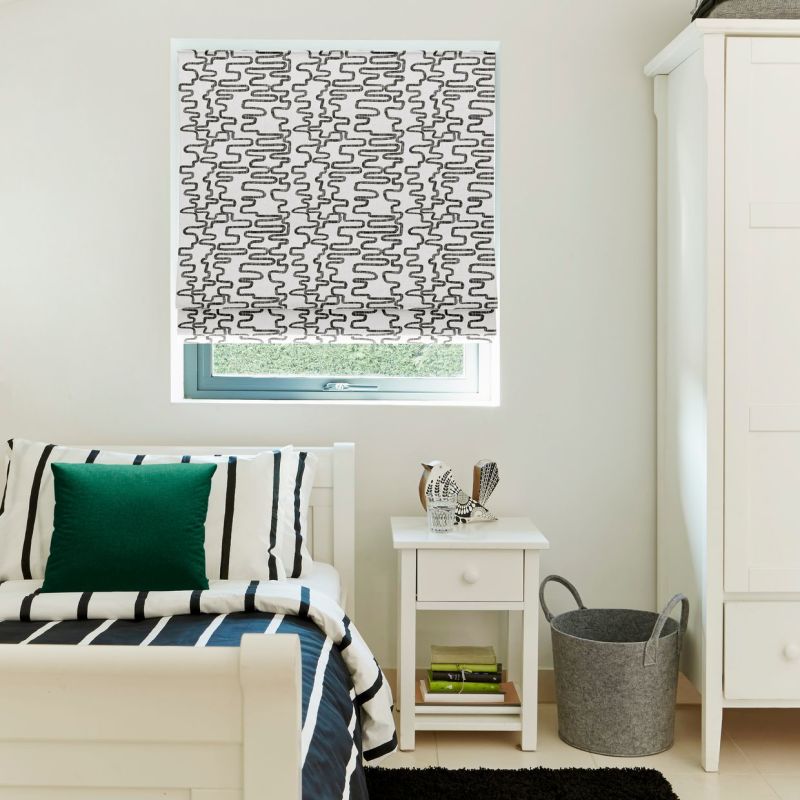 White roller blind with black abstract squiggle pattern lowered over window, filtering daylight; white bed with black-striped duvet and green cushion beside a small white nightstand; pale bedroom, greenery outside.