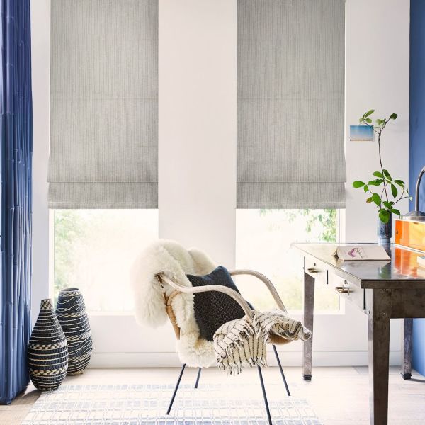 Grey fabric Roman shades, lowered across two tall windows with vertical texture; softly filtering daylight; bright home office corner with desk, sheepskin-draped chair, rug, blue side curtains and vases.