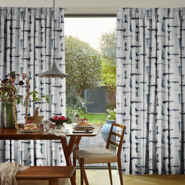 Floor-to-ceiling pleated curtains with a vertical black-blue watercolor brushstroke pattern, parted centrally to reveal a sunlit backyard; framing a dining table set for breakfast with wooden chairs and pendant lamp.