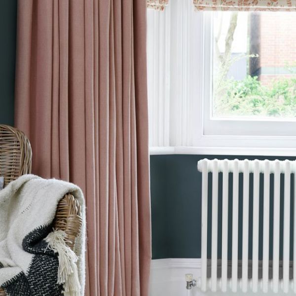 A floor-length dusty-rose curtain (left) and floral Roman blind are partially drawn, filtering soft daylight beside a window above a radiator in a cozy corner with a wicker chair.