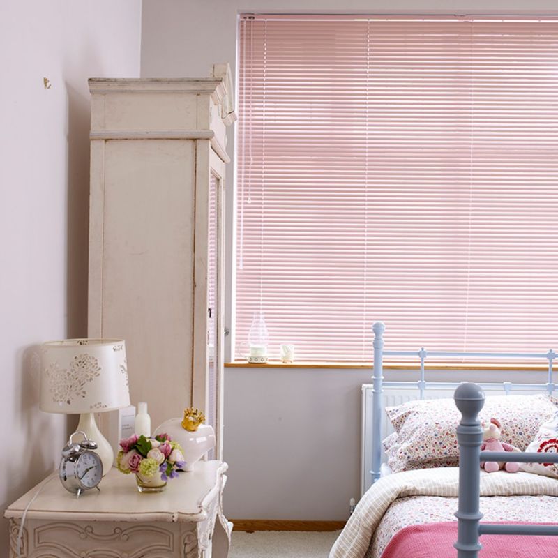 Pale pink horizontal blinds cover a wide window, closed and softly diffusing daylight; in a pastel bedroom with a light-blue metal bed, bedding, white nightstand, lamp and vase of roses.