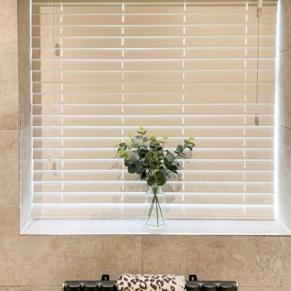 White horizontal blinds (Venetian), lowered and closed with side pull cords, filtering daylight; set in a tiled bathroom window recess with a glass vase of eucalyptus on the sill.