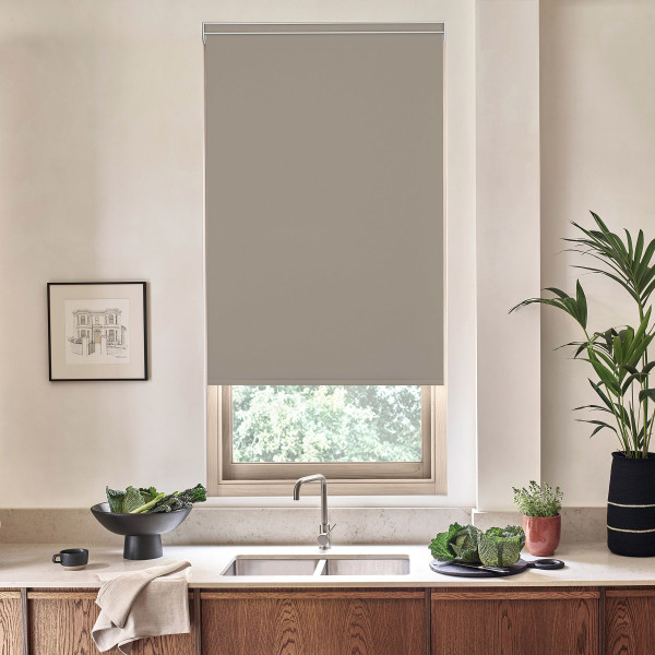 Roller blind lowered over a kitchen window, blocking light; stainless faucet and sink below on a stone countertop with wooden cabinets, potted plants, leafy vegetables, and framed sketch nearby.