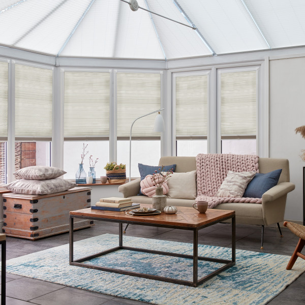 Pleated beige cellular blinds, lowered and softly diffusing daylight; bright conservatory-style living room with a neutral sofa, chunky pink throw, wooden coffee table, and patterned blue rug.