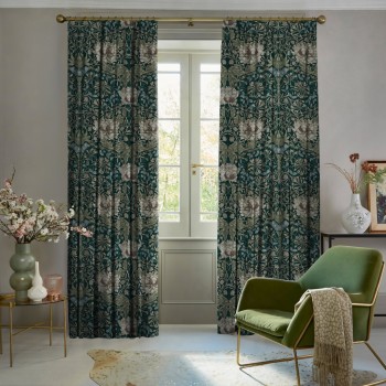 Heavy full-length floral curtains on a brass rod, teal background with cream flowers, parted to reveal a French door; filtering daylight into a cozy living room with a green armchair.