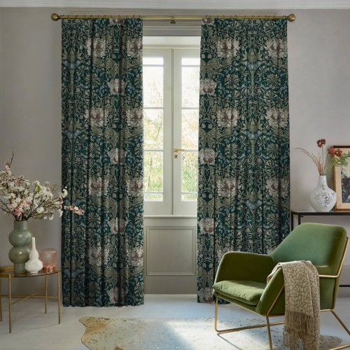 Heavy full-length floral curtains on a brass rod, teal background with cream flowers, parted to reveal a French door; filtering daylight into a cozy living room with a green armchair.