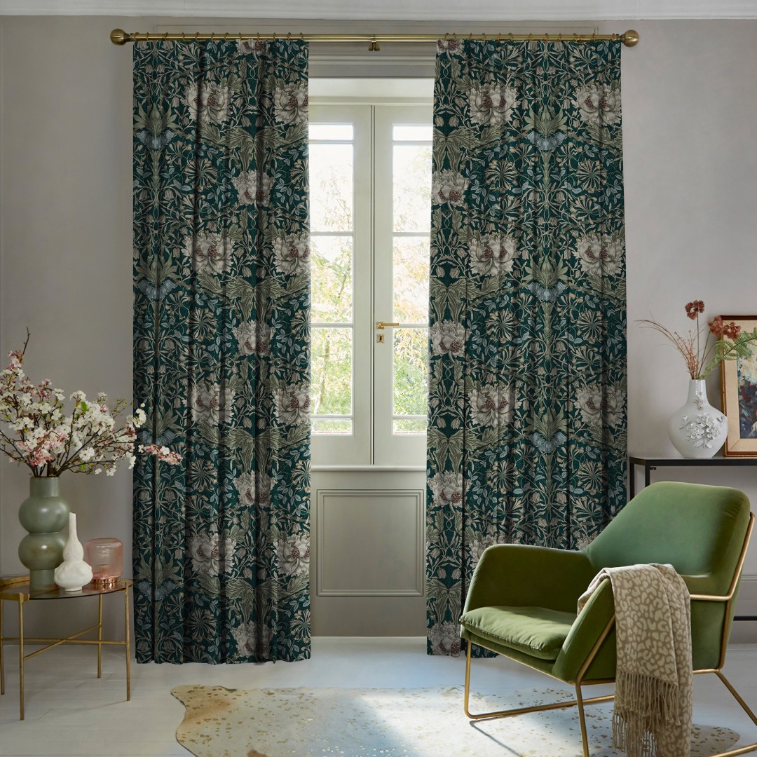 Heavy full-length floral curtains on a brass rod, teal background with cream flowers, parted to reveal a French door; filtering daylight into a cozy living room with a green armchair.