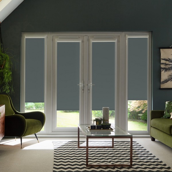 White French doors with gray roller blinds lowered halfway, filtering daylight into a modern living room framed by green velvet armchairs, glass coffee table atop zigzag rug, garden visible outside.