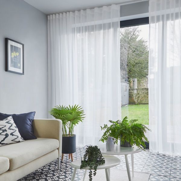 Sheer white floor-to-ceiling curtains on a ceiling track, softly filtering daylight and parted at a sliding glass door — living room with sofa, patterned tile floor, potted plants, garden view.