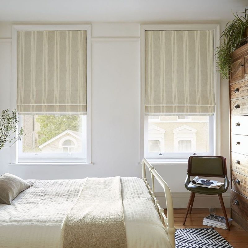Beige-striped Roman blinds, lowered and partially folded, filtering soft daylight; bedroom with cream metal bed, wooden chest of drawers, green chair, plants and a view of neighboring brick facades.
