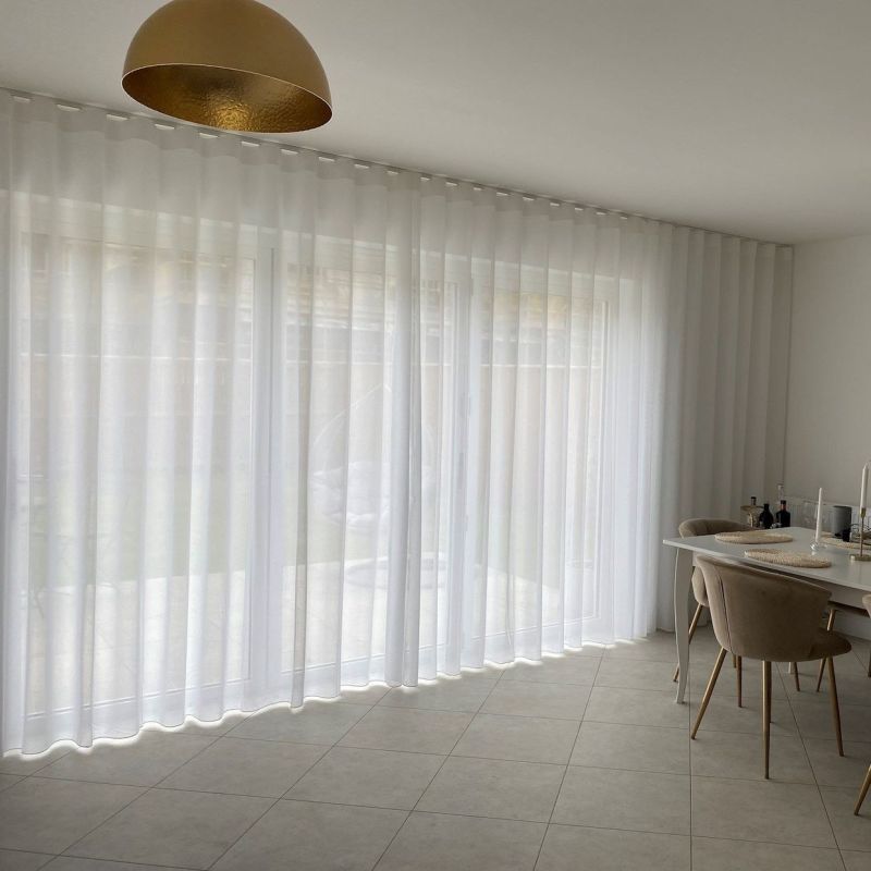 Sheer white floor-to-ceiling curtains on a ceiling track, forming soft vertical waves across wide glass doors, gently diffusing daylight into minimalist tiled dining area with beige chairs and gold pendant.