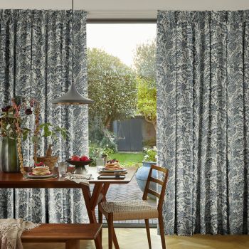 Floor-length pleated curtains with blue botanical print, mostly closed but parted to reveal a glass door; filtering sunlight into a dining area with wooden table set and sunlit garden beyond.