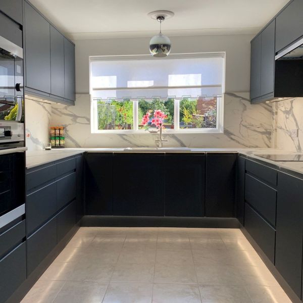 White roller blind with scalloped lower edge, lowered across a wide window, filtering daylight; modern U-shaped kitchen with dark cabinets, marble splashback, central sink and garden view with pink flowers.