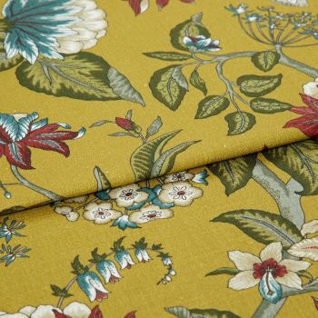Mustard-yellow fabric with botanical green leaves and red-and-blue flowers, folded once with overlapping edge; close-up studio-lit view of the textile on a flat surface.