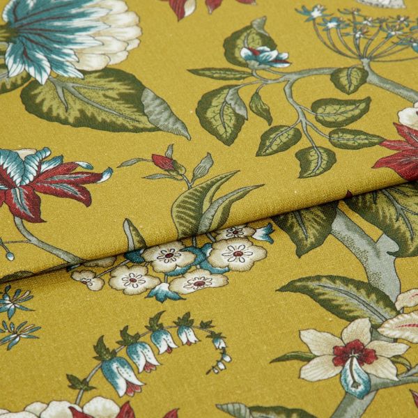 Mustard-yellow fabric with botanical green leaves and red-and-blue flowers, folded once with overlapping edge; close-up studio-lit view of the textile on a flat surface.
