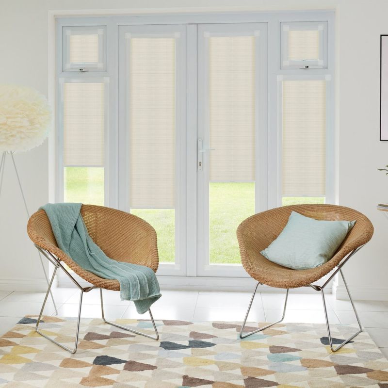 Beige roller blinds fitted inside white French doors and sidelights, lowered to partially filter daylight; bright living room with two wicker chairs, pastel throws, patterned rug and lawn view.