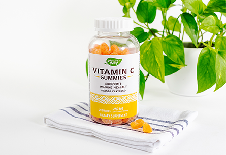 Nature's Way Vitamin C Gummies bottle with 3 gummies outside of the bottle sitting on a kitchen towel on a white surface with Pothos plant in the background 