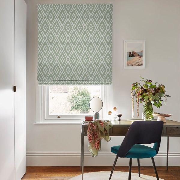 Roman blind with green-and-white diamond pattern lowered over a window, softly filtering daylight; desk with mirror, teal chair, vase of foliage and framed print in a home office.