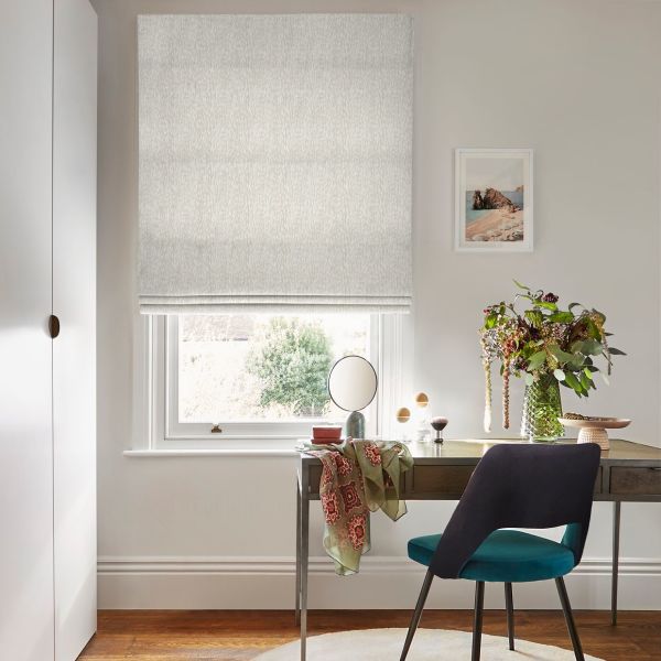 A light-gray textured Roman blind, lowered to just above the sill and softly filtering daylight, covers a window above a desk with mirror, vase of flowers and a teal chair.