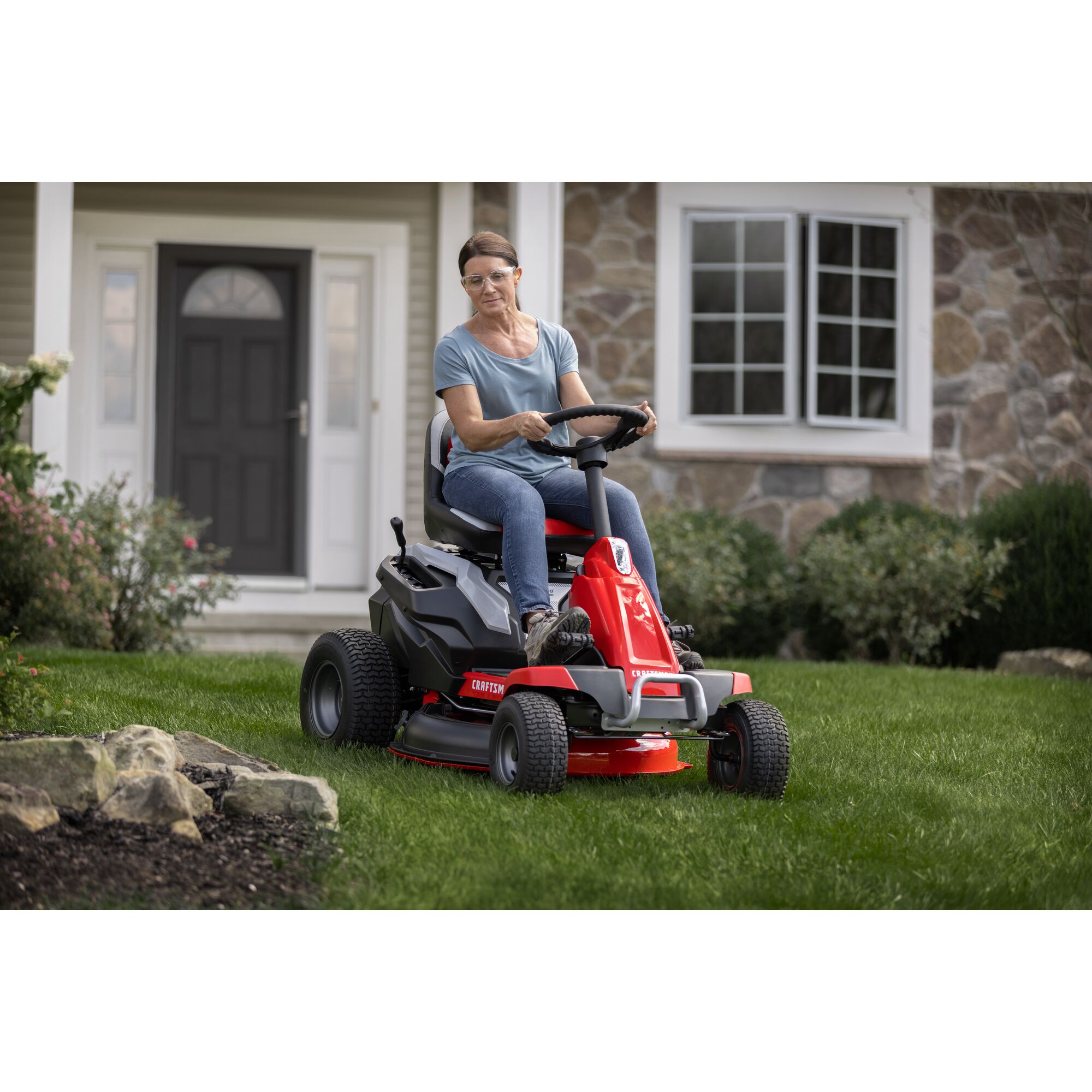 CRAFTSMAN Battery-Powered Compact Mower cutting grass around flowerbed with house in background