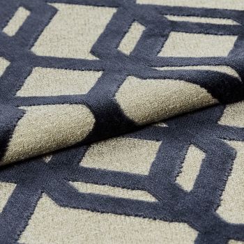 Beige woven rug with raised navy geometric lattice pattern â€” folded and layered to display plush pile â€” close-up on a neutral indoor surface under soft, directional lighting.