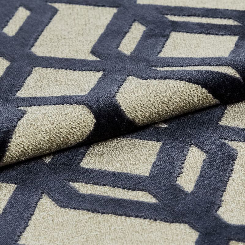 Beige woven rug with raised navy geometric lattice pattern — folded and layered to display plush pile — close-up on a neutral indoor surface under soft, directional lighting.