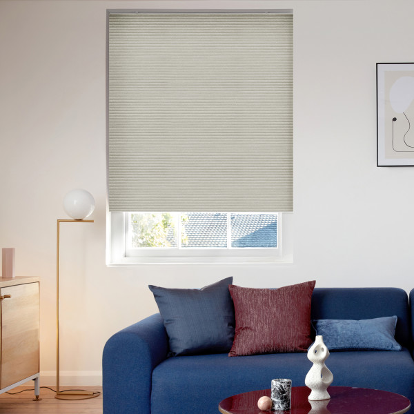 Beige cellular blind fully lowered over window, textured ribs; filtering daylight and covering most glass; modern living room with blue sofa, cushions, floor lamp, and a rooftop view outside.