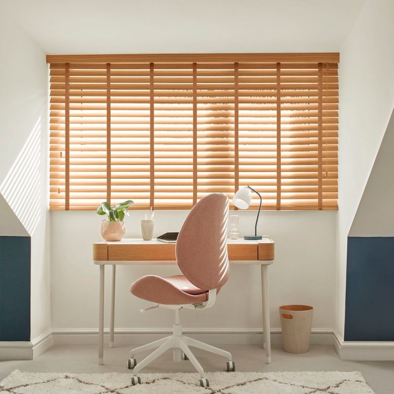 Wooden horizontal slat blinds covering a wide dormer window, partially closed and filtering warm sunlight; small home office desk with pink swivel chair, lamp, plant, waste bin and light carpet.
