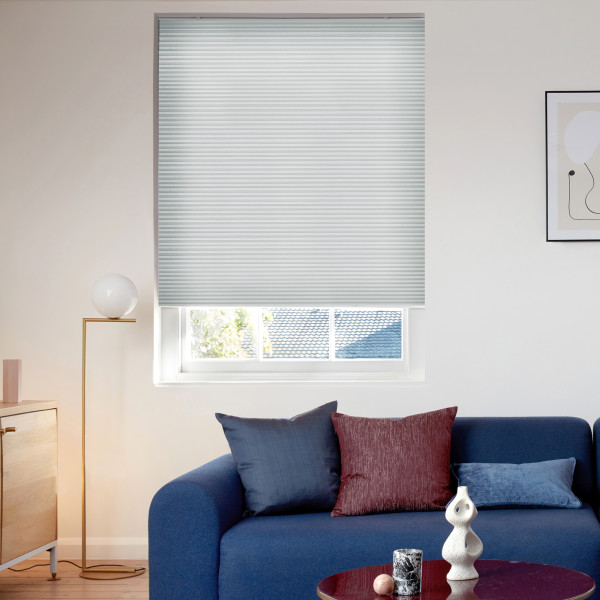 Light gray honeycomb pleated cellular blind lowered over a recessed window, filtering daylight; living room context with a blue sofa, colored cushions, floor lamp, framed abstract art and small coffee table.