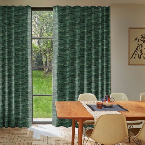 Green patterned, floor-length pleated curtains, mostly closed with a narrow central parting, filtering daylight and revealing a grassy backyard tree; mid-century dining room table, beige chairs and teapot nearby.